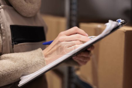 Photo for Postal office storage worker holding clipboard in warehouse, marking inventory checklist. Warehouse employee hand writing with pen close up, checking cardboard boxes in storehouse - Royalty Free Image