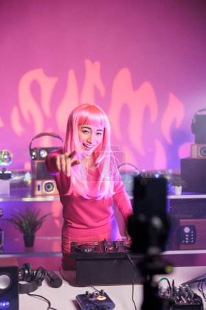 Photo for Woman with pink hair playing techno song at professional turntables while filimg process with mobile phone camera. Artist doing performance at nightclub with professional audio equipment - Royalty Free Image