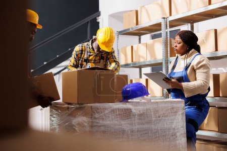 Photo for African american warehouse manager controlling assistant packing cardboard box. Supervisor holding clipboard with instruction and watching storehouse worker sealing cardboard box with adhesive - Royalty Free Image