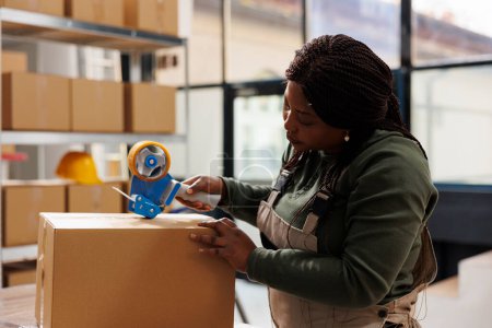 Photo for African american worker preparing customers orders, putting adhesive tape on cardboard box before shipping packages. Small business employee working at goods inventory in storage rooom - Royalty Free Image