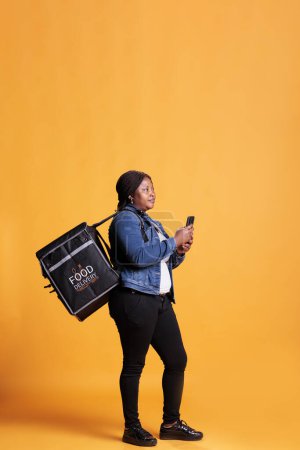Téléchargez les photos : Afro-américaine livreuse tenant téléphone en regardant adreess client sur l'application de restauration rapide avant de commencer à livrer ordre à emporter. Pizzeria courier portant sac à dos thermique à emporter en studio - en image libre de droit