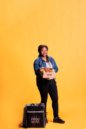 Foto de Distribuidora afroamericana sosteniendo caja llena de verduras saludables lista para entregar al cliente para el almuerzo fresco orgánico. Un mensajero sonriente llevando una mochila para llevar en el estudio - Imagen libre de derechos