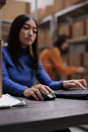 Photo for Asian warehouse operator coordinating order shipping on laptop. Storehouse young woman employee managing freight distribution and supply chain operations on computer in storage room - Royalty Free Image