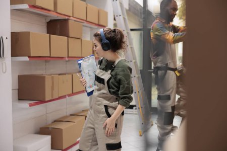 Photo for Warehouse employee working at products delivery, looking at shelves full with cardboard boxes. Storage room supervisor listening music while checking customers shipping details in storehouse - Royalty Free Image