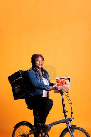 Téléchargez les photos : Livreuse d'épicerie afro-américaine à vélo tout en livrant une boîte remplie de légumes biologiques au client pour un déjeuner sain tout en utilisant le vélo comme moyen de transport. Emporter service alimentaire et concept - en image libre de droit