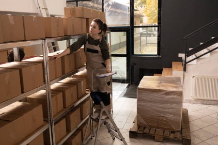 Photo for Storage room employee checking cardboard boxes barcode, standing on ladder in warehouse. Woman manager preparing orders for clients before start shipping packages in storehouse - Royalty Free Image