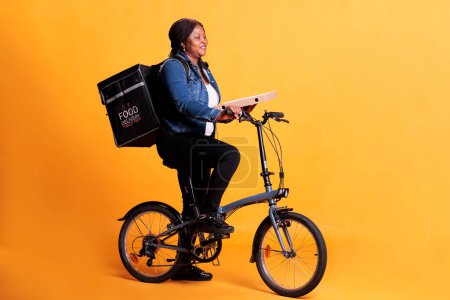 Téléchargez les photos : Courrier pizzeria afro-américain transportant une boîte de carton de pizza livrant une commande à emporter au client pendant l'heure du déjeuner, en utilisant le vélo comme moyen de transport en studio. Emporter service alimentaire et concept - en image libre de droit