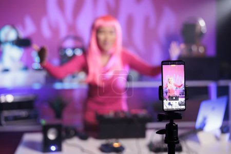 Téléchargez les photos : Femme aux cheveux roses debout à la table dj mélangeant chanson et platines tournantes devant la caméra tout en enregistrant une vidéo avec téléphone. Artiste souriant faisant la performance à la boîte de nuit avec équipement audio - en image libre de droit