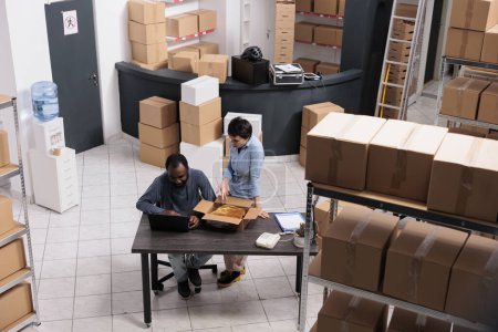 Photo for Top view of employees looking at cargo stock on laptop computer while putting trendy blouse in cardboard box preparing clients orders. Workers discussing shipment details working in warehouse - Royalty Free Image