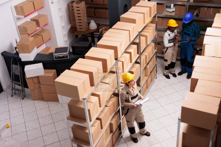 Photo for Diverse shipping service supervisors team working in warehouse, checking inventory system. Storehouse assistants reading stock supply report on clipboard and scanning parcels top view - Royalty Free Image