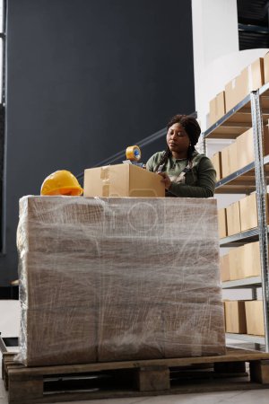 Téléchargez les photos : Gestionnaire afro-américain utilisant du ruban adhésif pour emballer les commandes des clients, préparer les colis pour la livraison dans la salle de stockage. Employé d'entrepôt portant l'ensemble industriel pendant l'inventaire de marchandises - en image libre de droit
