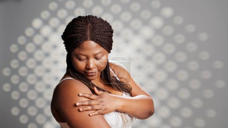 Photo for African american girl feeling radiant applying cream in studio, promoting skincare products for cosmetics campaign. Flawless woman using moisturizer or serum on camera, self love. - Royalty Free Image