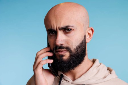 Foto de Hombre árabe confuso respondiendo a la llamada del teléfono inteligente y frunciendo el ceño retrato de las cejas. Joven barbudo calvo hablando por teléfono móvil y mirando a la cámara con expresión incierta - Imagen libre de derechos