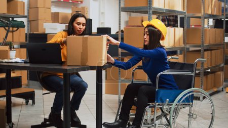 Foto de Mujer asiática en silla de ruedas llevando cajas de cartón para hacer control de calidad, trabajando con empleados. Trabajadora mirando mercancía en almacén, gestión de la cadena de suministro. - Imagen libre de derechos