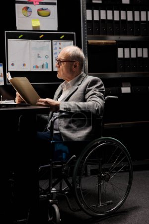 Téléchargez les photos : Homme d'affaires cadre supérieur en fauteuil roulant vérifier les documents de données du plan budgétaire de comptabilité. Employé bureaucratique expérimenté dans le dépôt rempli de dossiers de documents rapports de graphiques - en image libre de droit