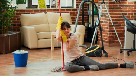 Photo for Exhausted woman sitting on living room floors with mop and bucket, feeling satisfied about finishing spring cleaning session. Young person being tired after tidying up household, doing chores. - Royalty Free Image