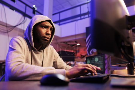 Foto de Interesado hacker cracking contraseña y la violación en el sistema de red informática. Hombre afroamericano concentrado en el capó que se dedica a la actividad criminal en línea y violar la ley - Imagen libre de derechos
