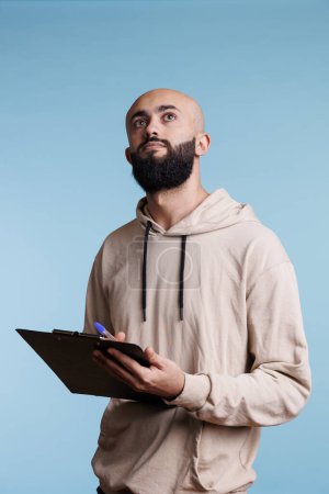 Foto de Hombre árabe pensativo sosteniendo portapapeles y contemplando la lista de verificación mientras mira hacia arriba con expresión facial reflexiva. Joven pensando mientras toma notas y planea - Imagen libre de derechos