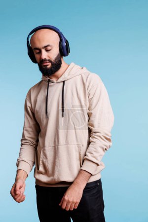 Téléchargez les photos : Homme arabe debout dans la pose détendue et écouter des chansons playlist dans les écouteurs. Jeune Arabe en sweat à capuche jouissant de musique, portant des écouteurs sans fil et regardant vers le bas - en image libre de droit