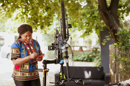 Photo for Enthusiastic african amercian female cyclist carefully examining specialized tools for servicing bicycle outside. Sporty black woman checking gear in home yard, dedicated to summer bike maintenance. - Royalty Free Image