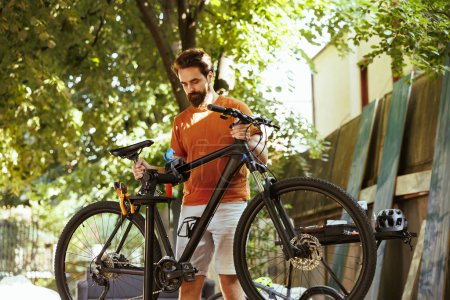 Photo for Sporty youthful man repairs bicycle damages in backyard using professional repair-stand and tools. Active young caucasian male carefully carrying and securing bike for maintenance. - Royalty Free Image