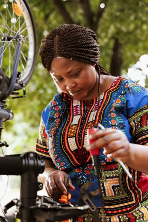 Foto de Mujer negra amante de los deportes que sirve y ajusta los componentes de la bicicleta con herramientas de trabajo expertas para el mantenimiento anual de verano. Saludable señora afroamericana examinando kit de herramientas y reparación de bicicletas. - Imagen libre de derechos
