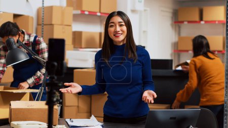 Photo for Business owner filming teleshopping video on camera, advertising merchandise in storage room. Young woman using social media streaming platform to create online promotion, marketing. Tripod shot. - Royalty Free Image