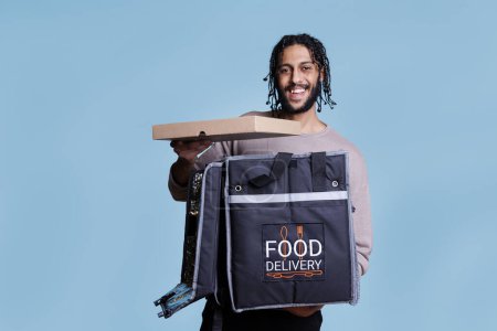 Photo for Happy arab courier holding pizza box and looking at camera with cheerful facial expression. Carefree pizzeria delivery service worker taking fast food from thermal backpack portrait - Royalty Free Image