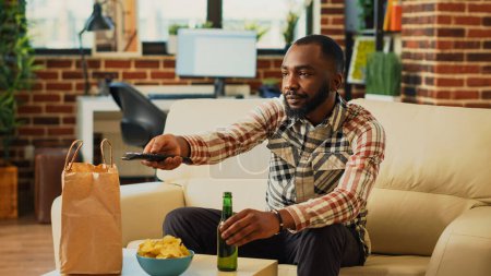Photo for Relaxed guy having meal from fast food takeout place, unpacking food from delivery bag in front of television. Young person preparing to eat dinner and watch comedy tv show or movie on sofa. - Royalty Free Image