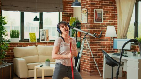 Téléchargez les photos : Positive woman listening to music on headphones and washing apartment floors, sweeping dirt. Housewife feeling happy cleaning household with mop and appliances, dance moves. - en image libre de droit