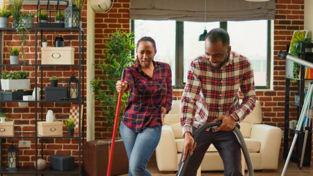 Foto de Happy man and woman showing dance moves and doing spring cleaning, having fun with music cleaning floors with mop and vacuum cleaner. Young smiling couple doing chores. Handheld shot. - Imagen libre de derechos