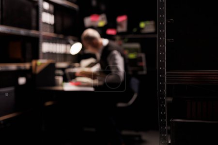 Photo for Selective focus of dark room equipped with metallic shelves full with confidential documents. In background old private detective working overhours at criminal case, analyzing investigation evidence - Royalty Free Image