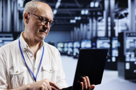 Photo for Senescent system administrator inspects server clusters in data center, ensuring smooth performance. Precise professional monitoring energy consumption across units components using laptop - Royalty Free Image