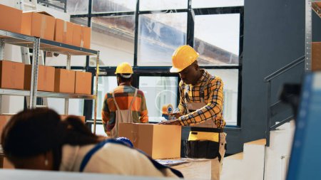 Téléchargez les photos : African american men packing products in boxes, using warehouse tools to plan retail shipment in storage room. Male employees working with products logistics, shipping merchandise. Handheld shot. - en image libre de droit