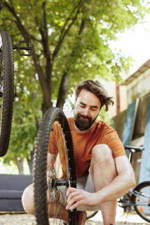 Photo for Active and committed man crouching to repair bike wheel rim in home yard using professional tool. Healthy male cyclist working on dismantled bicycle tire outside using specialized wrench. - Royalty Free Image