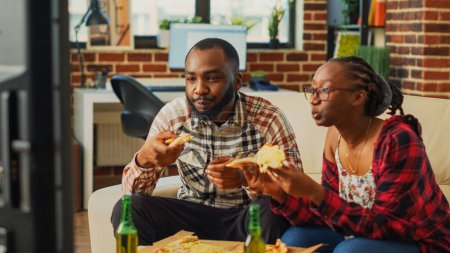 Téléchargez les photos : Happy life partners eating slices of pizza on couch, having fun together watching favorite movie on television. Young man and woman in relationship enjoying delivery food and alcohol. - en image libre de droit