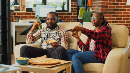 Téléchargez les photos : Partenaires afro-américains mangeant des tranches de pizza à la maison, se sentant heureux de regarder le film ensemble. Couple détendu s'amusant à manger à emporter livraison de nourriture dans le salon, activité de loisirs. - en image libre de droit