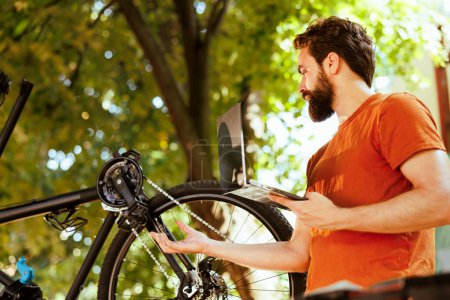 Photo for In his yard, active sports-loving man uses smart digital device to investigate bicycle repair possibilities. Caucasian male cyclist inspects damaged bike with laptop outdoors. - Royalty Free Image