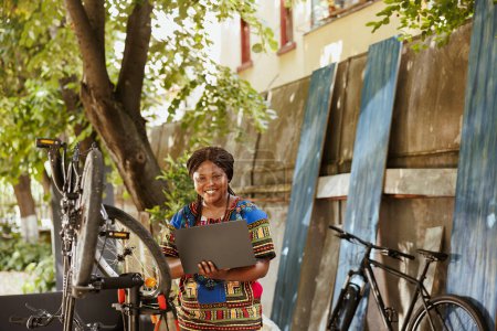 Foto de Entusiasta mujer afroamericana amante de los deportes con portátil reparación de bicicletas en el patio de casa. Joven atlética mujer negra que utiliza minicomputador para la investigación de mantenimiento de bicicletas. - Imagen libre de derechos