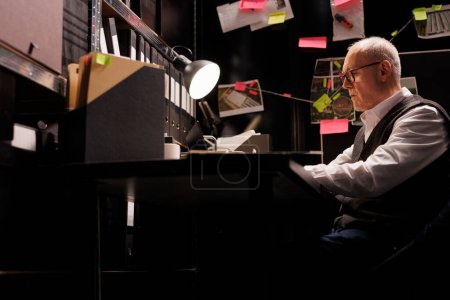 Photo for Senior police officer analyzing federal documents, working overtime at criminal case in arhive room. Elderly private detective analyzing mysterious suspect report, checking victim files - Royalty Free Image