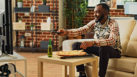Téléchargez les photos : African american guy enjoying pizza from delivery, binge watching favorite tv show in living room. Young happy man eating fast food from takeaway place, sitting at home with action film. - en image libre de droit