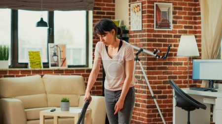 Téléchargez les photos : Modern person using cleaning appliances to sweep floors, vacuuming in living room. Young casual woman picking up dust and dirt with vacuum cleaner, using washing solution and all purpose cleaner. - en image libre de droit
