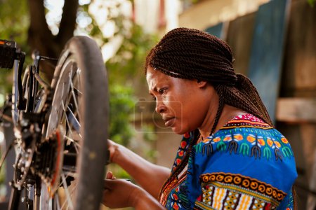 Téléchargez les photos : Image détaillée du vélo de réparation afro-américain féminin pour un vélo de plein air sécurisé. Femme noire cycliste en bonne santé analyse étroitement la chaîne de vélo en utilisant un outil professionnel pour maintenir l'efficacité du cycle. - en image libre de droit
