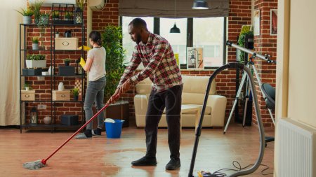 Téléchargez les photos : Interracial couple vacuuming floors and cleaning furniture on shelves, sweeping dust or dirt with tools. Life partners using vacuum cleaner with hose, gloves and rags to clean household. House chores. - en image libre de droit