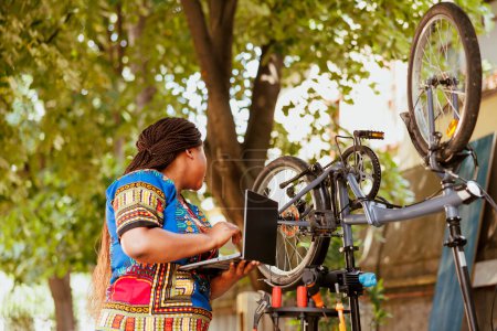 Foto de Saludable y vibrante fermale afroamericano que investiga en Internet para el mantenimiento de bicicletas para el ciclismo de ocio al aire libre. Mujer negra amante de los deportes revisando equipos de bicicleta en el portátil. - Imagen libre de derechos