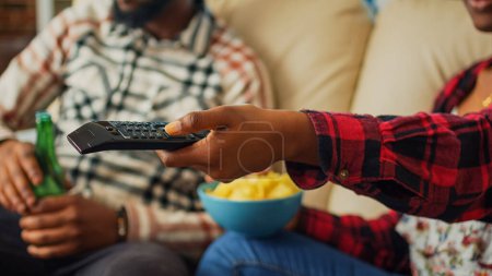 Photo for Girlfriend switching channels on television to find movie, using tv remote control to watch favorite film or show with man. Young couple eating chips and drinking beer at home. Close up. - Royalty Free Image