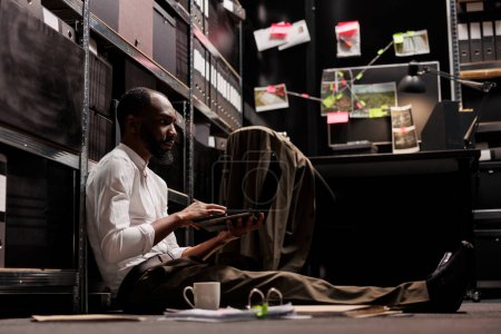 Foto de Policía afroamericano sentado en el suelo y trabajando en la computadora portátil en la oficina por la noche. Investigador de la policía examinando pistas y fotos sospechosas en la computadora para resolver el caso de crimen - Imagen libre de derechos