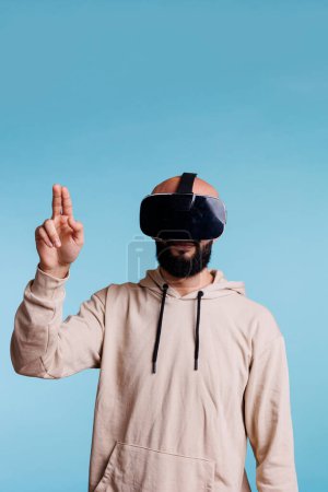 Téléchargez les photos : Homme portant vr siège de tête tout en jouant jeu de réalité virtuelle et geste avec deux doigts. Jeune profitant de métaverse et expérience d'exploration de simulation cyberespace - en image libre de droit