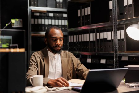 Téléchargez les photos : Un flic afro-américain qui fait des heures sup sur un ordinateur portable dans un bureau de détective sombre. Enquêteur sérieux analysant le rapport de police et cherchant des informations pour résoudre le crime la nuit - en image libre de droit
