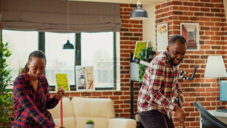 Foto de African american couple having fun spring cleaning with mop and vacuum cleaner, dancing and listening to music at home. Cheerful life partners mopping and vacuuming floors in household. - Imagen libre de derechos